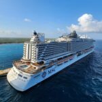 MSC luxury cruise ship sailing on deep blue ocean waters near a tropical coastline under a clear sky.