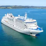Luxury cruise ship sailing across calm blue ocean waters with a scenic coastline in the background.